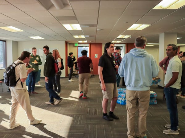 A group of people are socializing and eating in a large, brightly lit office environment during a casual gathering.