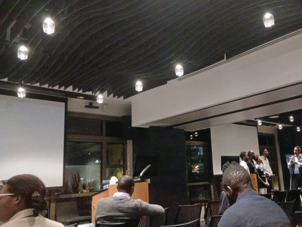 A photograph of a presentation or panel discussion inside a modern hall with an audience watching speakers on stage.