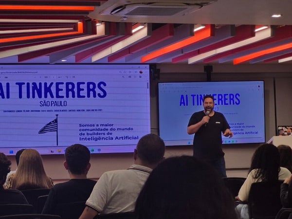 A man is giving a presentation at an 'AI Tinkerers' event in São Paulo, standing in front of large digital screens with an audience in the foreground.