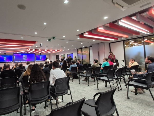 A photograph showing an audience of people seated in a modern conference room, watching a presentation displayed on multiple large screens.