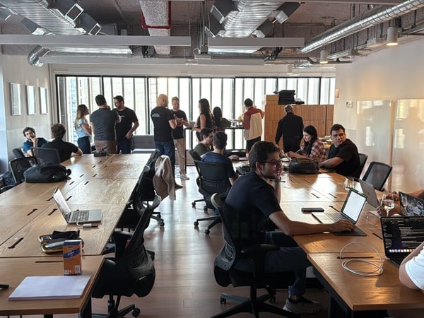 A candid photograph captures a large group of people working and socializing in a modern, open-plan office space featuring long wooden tables and exposed industrial ceilings.