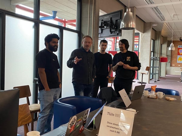 A group of four men are standing and talking in a modern indoor setting, likely a university or office, around a table with laptops and snacks.