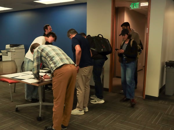 A group of people in an office setting, with one person signing papers at a table while others stand nearby or wait in a doorway.