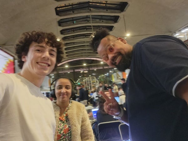 A group selfie taken indoors at a large, crowded event space, featuring three smiling people in the foreground. The man on the right is leaning in and making a peace sign gesture.