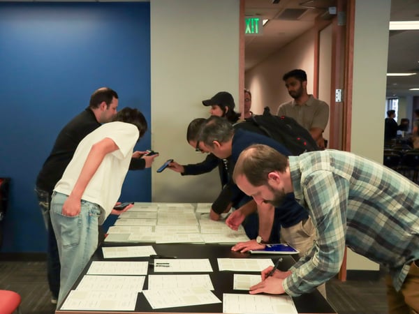 A group of people are gathered around a table covered in papers, with some individuals writing on them while others use their smartphones to take photos of the documents.