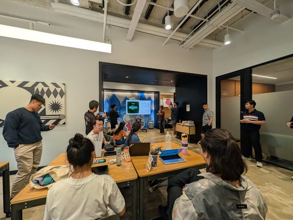 A candid photograph taken inside a modern office space showing a group of people gathered around desks and a doorway, where a presentation is being displayed on a screen in the background.