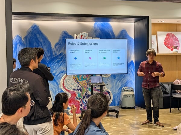A photograph capturing an indoor event where a man in a red plaid shirt is giving a presentation to a group of attendees, standing next to a large screen displaying rules and submission guidelines against a backdrop of a colorful blue and red mural.