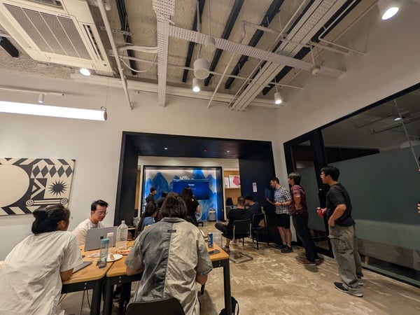 A casual gathering of people is taking place in a modern office space featuring exposed ceilings and concrete floors. Some attendees are seated at a table with food and laptops, while others stand near a doorway leading to a brightly lit presentation area.