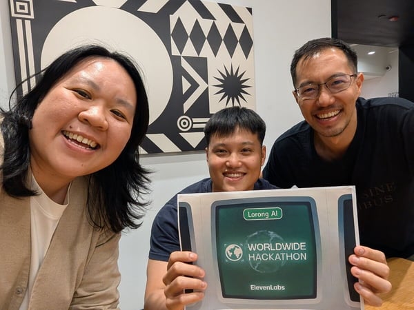 Three smiling individuals, one woman and two men, pose for a close-up group photo while holding a sign promoting the Lorong AI Worldwide Hackathon by ElevenLabs.