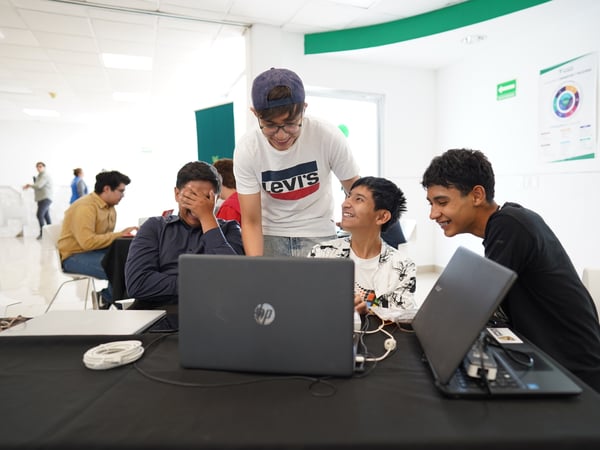 A group of young men are smiling and laughing while gathered around laptops in a bright, modern indoor space.