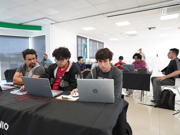 A group of young men are seated at tables in a brightly lit, modern room, focused on their laptops during what appears to be a workshop or hackathon.