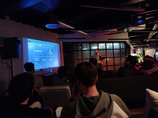 A low-light photograph captures an indoor presentation event where a speaker stands near a podium with a laptop, addressing an audience seated in the foreground, with a large screen displaying a blue-themed dashboard interface.