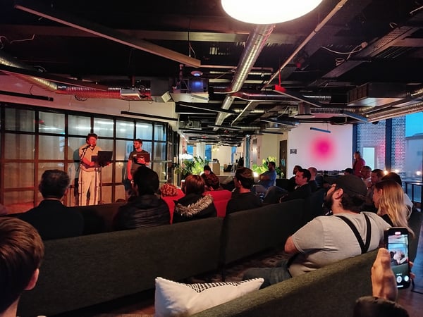 A photograph captures an evening event or presentation taking place in a dimly lit, modern venue with industrial design elements and exposed ceilings. Two presenters stand at the front addressing a large audience seated on long couches.