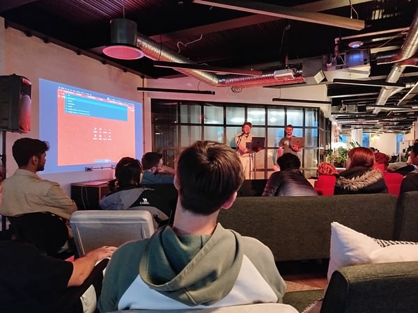 A candid photograph capturing an indoor tech presentation or meetup where two presenters are standing with laptops in front of a large projection screen displaying a software interface, while an audience watches from comfortable seating.