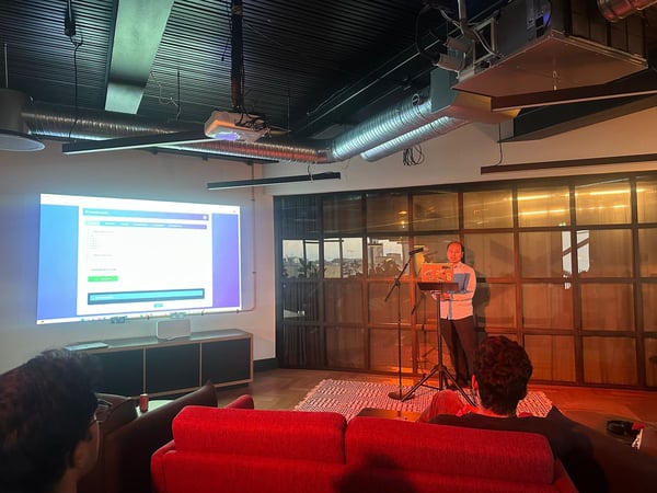 A photograph capturing an indoor presentation or tech talk, showing a speaker standing near a microphone and a projected screen displaying a web interface, while audience members sit on red couches in the foreground.