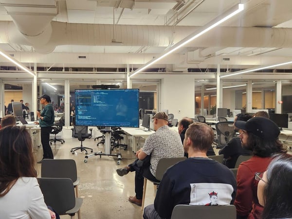 A photograph capturing a technical presentation or meetup held in a modern office space, showing an audience seated and facing a large screen displaying lines of code, with a speaker standing next to the display.