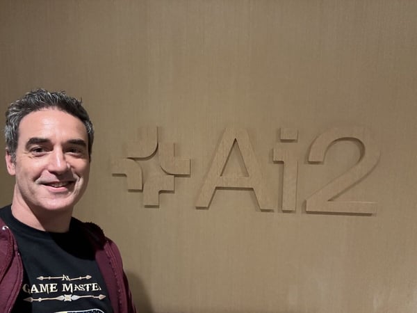 A man wearing a burgundy hooded sweatshirt and a black t-shirt smiles while taking a close-up selfie next to a wall featuring the wooden logo and text "+ Ai2".