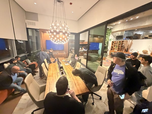 A candid photograph captures a group of approximately ten people gathered around a long, rustic wooden conference table in a modern, well-lit indoor meeting space.
