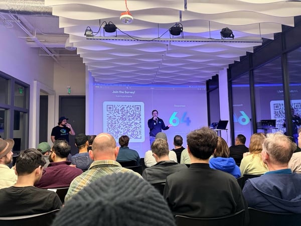 A photograph captures an indoor presentation where an audience is seated, facing a speaker standing in front of a large screen displaying a QR code for a survey and the current response count of '64'.