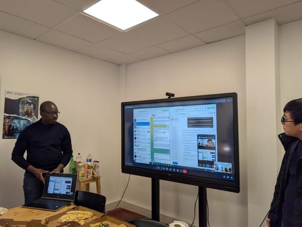 A man stands in an office setting next to a table with pizza and a laptop, observing a large screen that displays a video call and messaging interface.