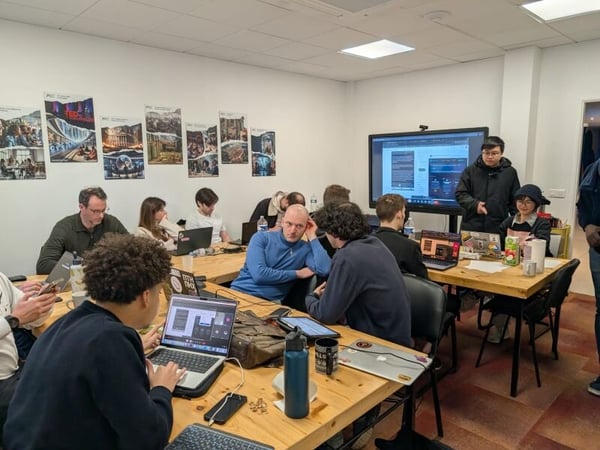 A group of people are gathered in a modern office or classroom setting, engaged in a collaborative activity with laptops and a large digital screen.