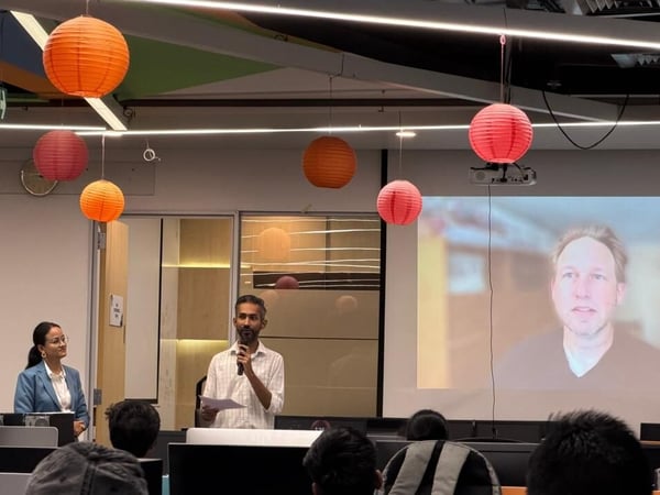 A man is giving a presentation in an office setting decorated with colorful paper lanterns, while a woman stands nearby and a remote participant is visible on a large screen.