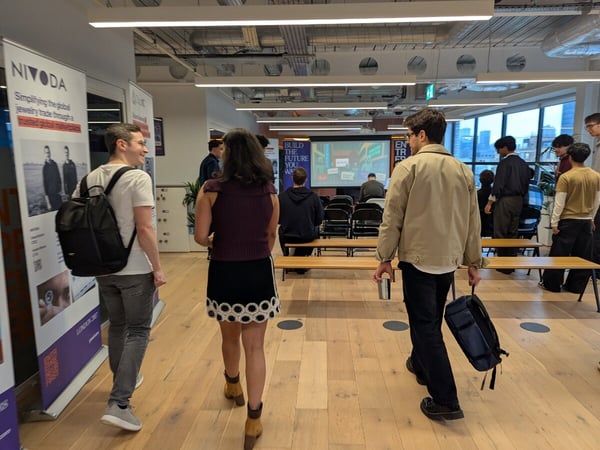 A candid photograph taken indoors at a modern event or conference venue, showing several attendees walking across a light wooden floor past event banners and toward a presentation screen in the background.