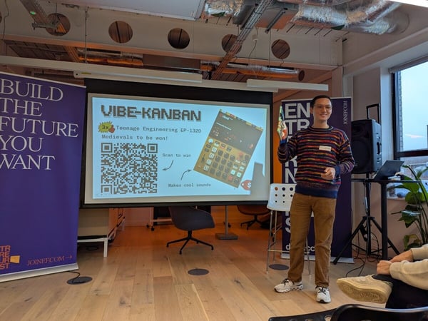 A man wearing a multicolored striped sweater gives a presentation in a modern event space, standing in front of a projection screen that displays a QR code and promotional text for a product called VIBE-KANBAN.