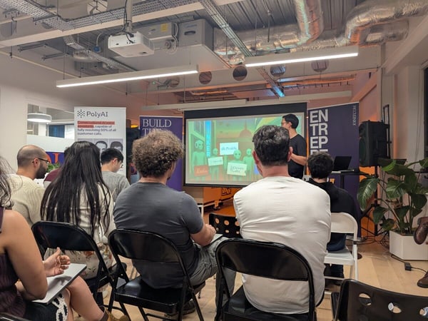 A photograph capturing an indoor technical presentation or meetup, showing an audience seated in folding chairs facing a projection screen and a speaker standing to the right.