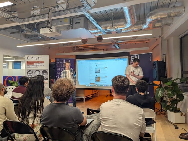 A candid photograph captures a technology event or presentation taking place indoors, showing two male presenters standing near a large projected screen while an audience watches from the foreground. The setting features exposed industrial ductwork on the ceiling and promotional banners for PolyAI and JOINF.COM.