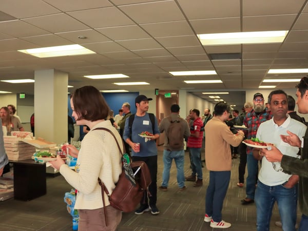 A group of people are gathered in an office setting for a social event, holding plates of food and engaging in conversation.