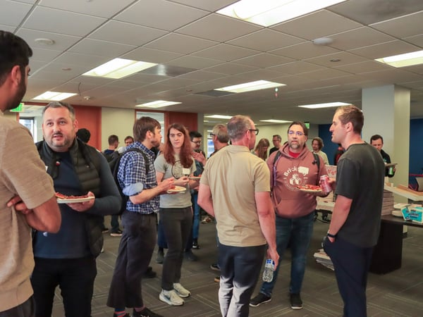 A group of people are gathered in an office setting, standing in small groups and talking while eating pizza.