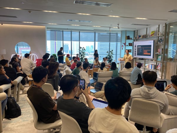 A group of people are gathered in a modern office space for a presentation. A speaker stands next to a large screen displaying an agenda while the audience sits on chairs and couches, some using laptops and phones.