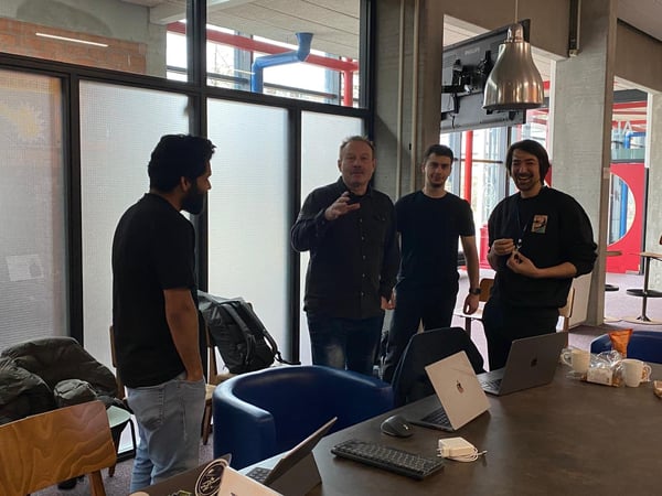 A group of four men are gathered around a table in a modern office or workshop space, engaged in a discussion or demonstration.