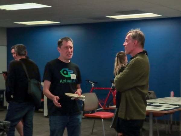 Two men are engaged in a conversation in an indoor setting, with one man wearing a black t-shirt and holding a plate while the other listens with his arms crossed.
