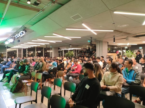 A candid photograph capturing a large audience seated in a modern, dimly lit indoor venue, attending an event or presentation.