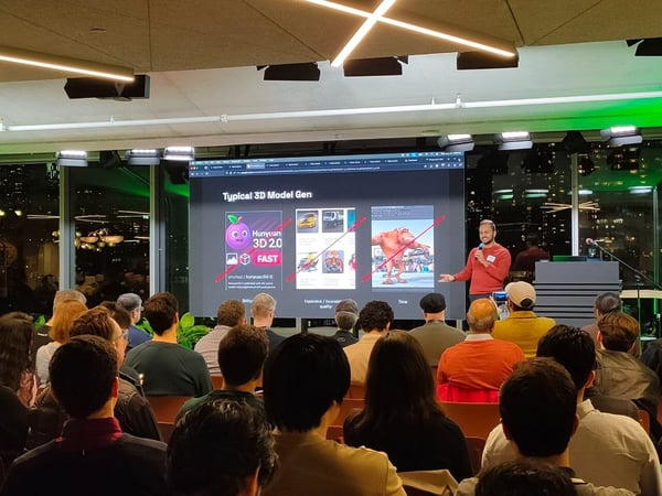 A speaker in a red shirt addresses a large, seated audience in a dimly lit, modern venue, presenting a slide about 3D model generation on a wide screen with a city skyline visible through the windows behind him.