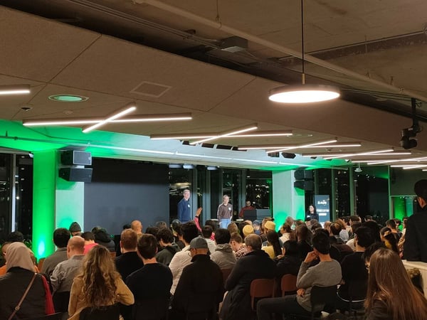 A photograph capturing a large indoor nighttime event where an audience is seated, facing a stage area illuminated by green accent lights, with two speakers standing at the front.