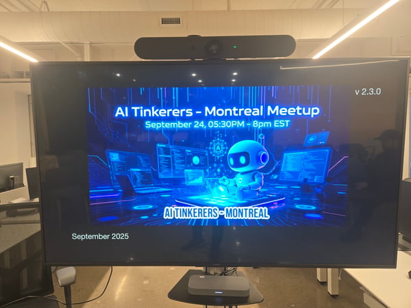 A photograph taken in an indoor office setting, showing a large flat-screen monitor displaying a futuristic, blue-themed slide for an event titled 'AI Tinkerers - Montreal Meetup.'