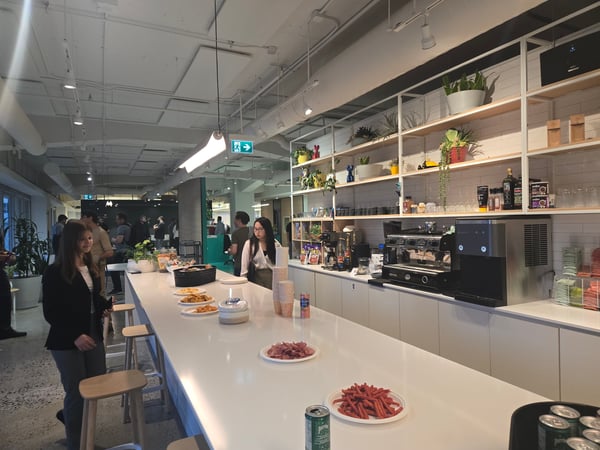 A candid photograph capturing a social gathering inside a modern office break room or event space, featuring a long white counter stocked with snacks and drinks, and several people mingling near a professional coffee station and shelving unit.