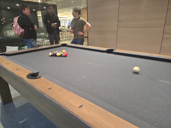 Three men are standing and conversing behind a large pool table set up with a racked set of balls and a cue ball in a modern, indoor recreational space.