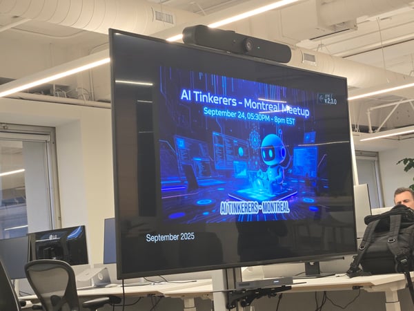 The image is a photograph taken inside a modern office, showing a large, vertically oriented screen displaying a promotional slide for the "AI Tinkerers - Montreal Meetup." The environment includes white walls, exposed ductwork, office desks, and a partially visible person seated on the right.