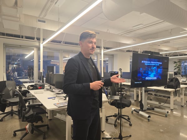 A man wearing a dark suit is giving a presentation in a modern, open-plan office space, gesturing toward a large television screen that displays the title slide for the 'AI Tinkers - Montreal Meetup.'