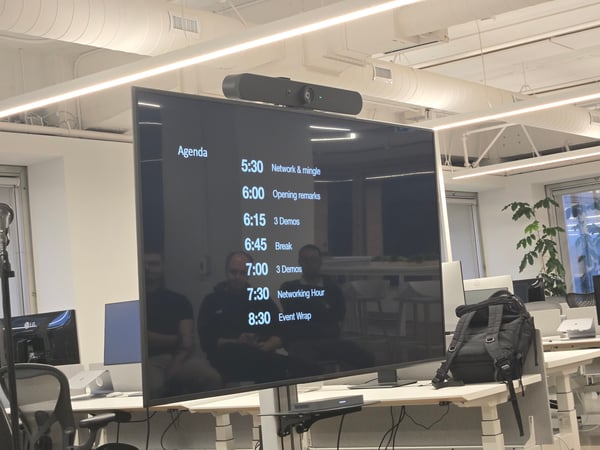 A photograph taken in a modern office space shows a large black monitor displaying an event agenda with times listed for networking, demos, and breaks. Reflections of people attending the event are visible on the dark screen surface.