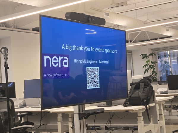 A photograph taken inside a modern office space, showing a large monitor displaying a presentation slide with a dark blue background that thanks event sponsors, features the 'nera' logo, and advertises a job opening for an ML Engineer in Montreal, along with a QR code.