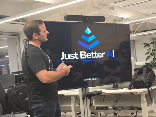 A man is giving a presentation in an office setting, standing next to a microphone while gesturing toward a large screen displaying the logo and text "Just Better AI" and "BUILDING AGENTS".
