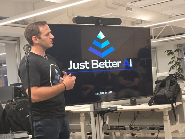 A man is giving a presentation or talk in a modern office setting, standing next to a large monitor that displays the logo and name of a company, "Just Better AI."