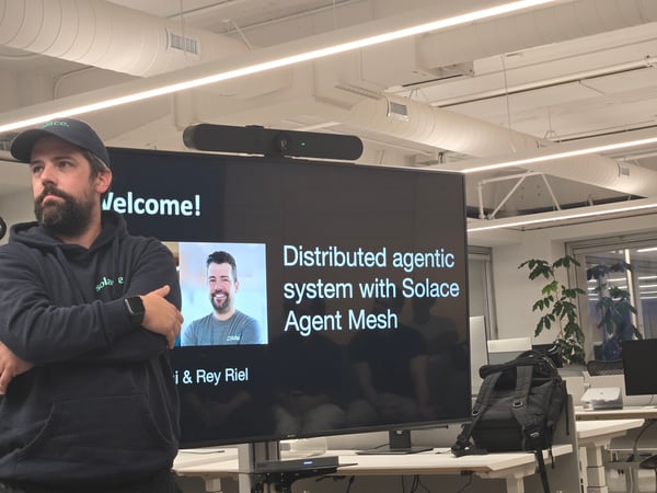 A photo taken during a technical presentation in a modern office, showing a bearded man standing in the foreground, partially obscuring a large screen displaying the title slide: 'Distributed agentic system with Solace Agent Mesh'.