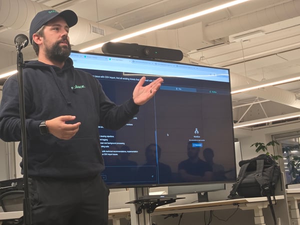 A bearded man wearing a dark hoodie and a baseball cap is giving a presentation or software demonstration in a modern office setting. He is standing next to a large monitor displaying a dark-themed application interface with workflow steps and technical notes.