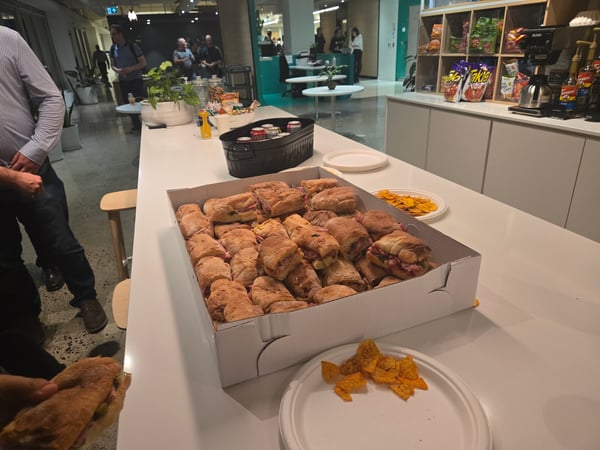 A candid photograph capturing a catering spread of sandwiches, chips, and canned drinks on a long white counter in a modern office break area, with several people socializing in the background hallway.